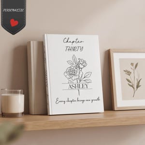 Może przedstawiać: Biała książka w twardej oprawie z czarną grafiką liniową przedstawiającą różę, napisem Chapter THIRTY, imieniem ASHLEY oraz frazą Every chapter brings new growth. Książka stoi obok zwykłej książki i oprawionego wydruku botanicznego.