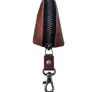 May include: A brown leather key pouch with a black zip and a metal clasp. The pouch is triangular and has a small leather strap attached to the clasp. The text "Handcrafted Leather Goods Etsy Shop - WildtharCO" is at the bottom.