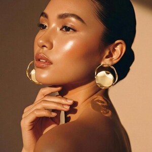 May include: A woman with radiant skin and dark hair, wearing large gold earrings. The earrings have a unique, organic shape. The woman's hand gently touches her neck. The lighting creates a warm, golden glow, highlighting her features and the jewelry.