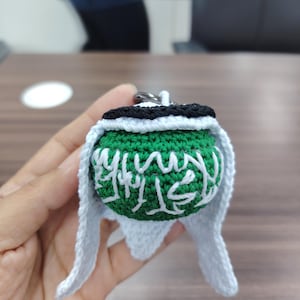 Könnte beinhalten: Ein handgefertigter Häkel-Schlüsselanhänger. Die grüne Kugel hat weiße arabische Kalligraphie, einen schwarz-weißen oberen Teil und weiße Seitenklappen. Der Artikel wird in der Hand einer Person gehalten.