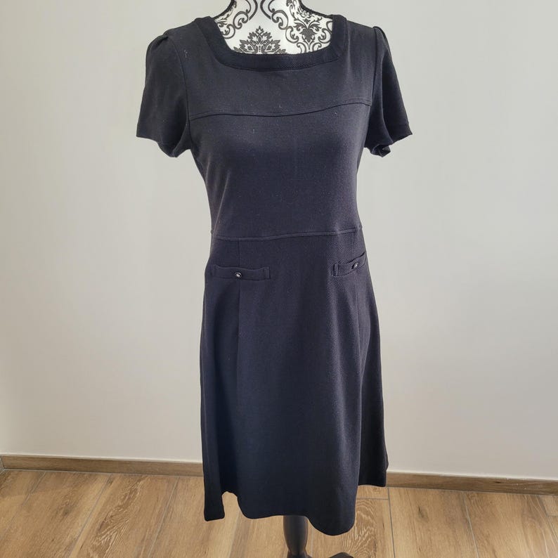 May include: A black short-sleeved dress with a square neckline and two small pockets. The dress has a fitted bodice and a slightly flared skirt. The dress is displayed on a mannequin.