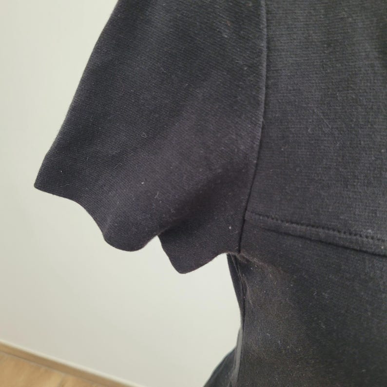 May include: Close-up of a black short-sleeved shirt. The sleeve has a scalloped edge. The fabric appears to be a knit material. The shirt is plain and has no visible patterns or embellishments.