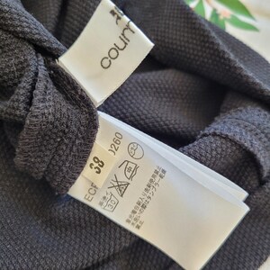 May include: Close-up of a dark gray garment with two attached white tags. One tag reads "courn" in a stylized font. The other tag displays care symbols and the number "38" and "0260".