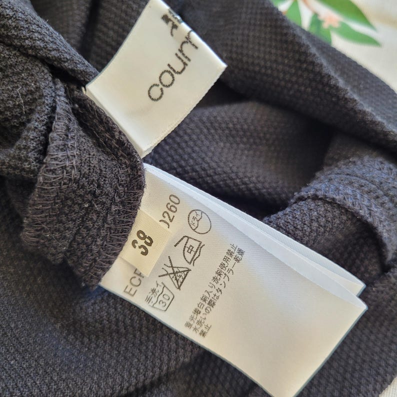 May include: Close-up of a dark gray garment with two attached white tags. One tag reads "courn" in a stylized font. The other tag displays care symbols and the number "38" and "0260".