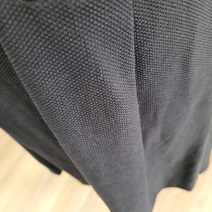 May include: Close-up of a black fabric with a textured, woven pattern. The material appears to be a knit or pique, with a slightly shiny appearance. The fabric is draped, showing folds and shadows.