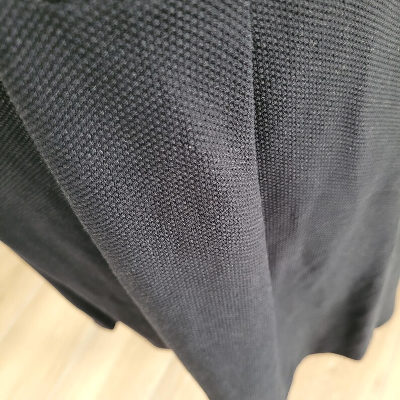 May include: Close-up of a black fabric with a textured, woven pattern. The material appears to be a knit or pique, with a slightly shiny appearance. The fabric is draped, showing folds and shadows.