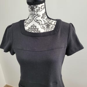May include: A black dress with short sleeves and a square neckline. The dress has a textured band across the chest and a textured neckline. The dress is displayed on a mannequin with a black and white patterned design.