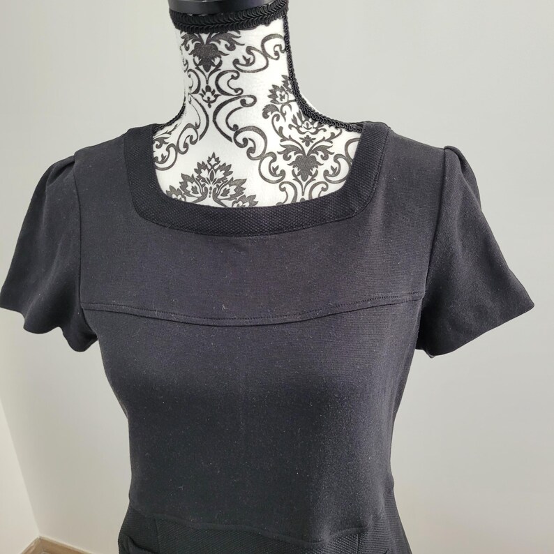 May include: A black dress with short sleeves and a square neckline. The dress has a textured band across the chest and a textured neckline. The dress is displayed on a mannequin with a black and white patterned design.