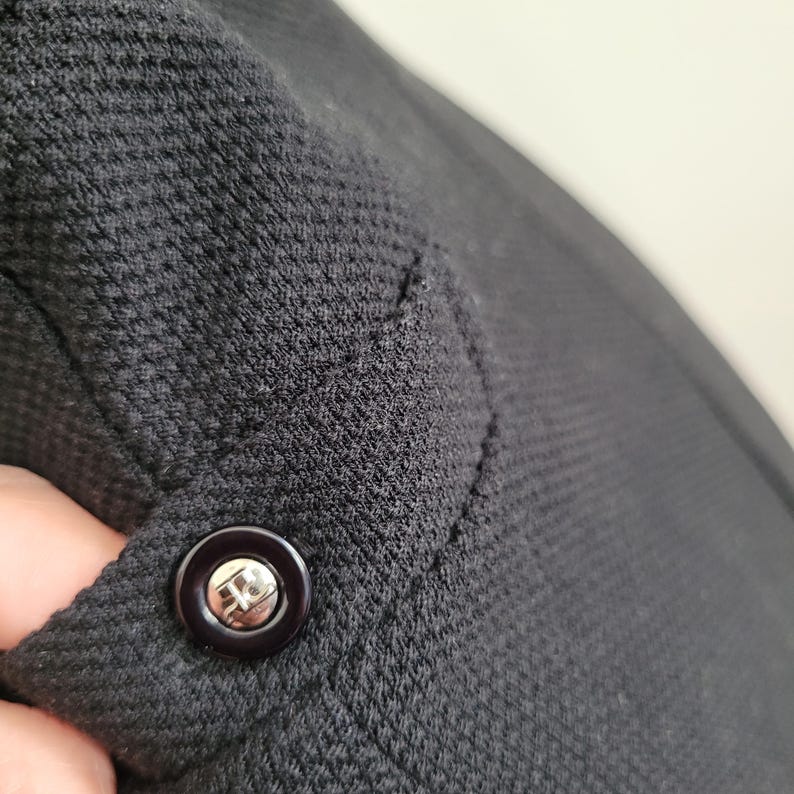 May include: Close-up of a black garment with a textured knit fabric. A dark button with a silver emblem is visible on the garment's edge. The fabric has a subtle pattern and a tailored seam.