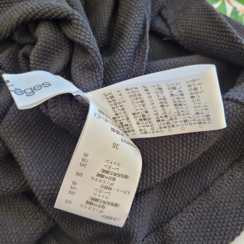 May include: Close-up of a dark gray knit garment with a white tag. The tag displays Japanese text and fabric composition percentages. The garment has a textured appearance and a label with the brand name "r&egrave;ges".