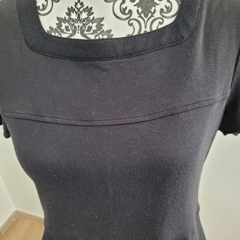 May include: A black dress with a square neckline and short sleeves. The neckline features a textured trim, and a horizontal seam detail is present across the chest. The dress is made of a soft, textured fabric.