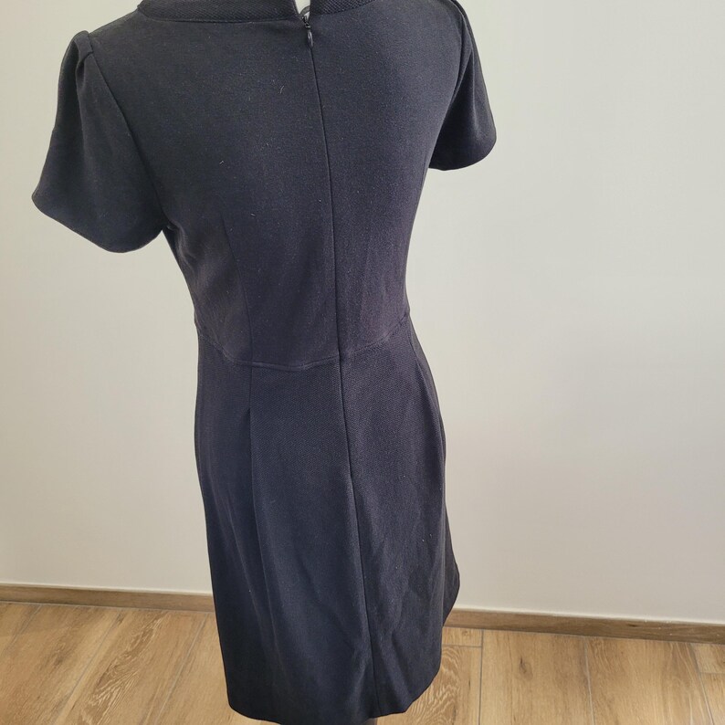 May include: A black dress with short sleeves and a knee-length hem. The dress has a fitted bodice and a slight A-line skirt. The back of the dress features a zipper closure and a small slit at the neckline. The dress is made of a solid, dark fabric.