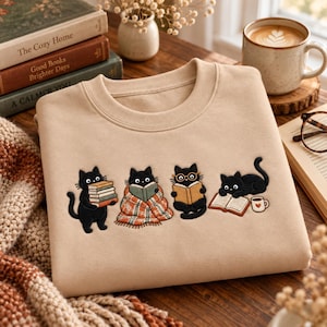Puede incluir: Una sudadera beige de cuello redondo con un bordado de cuatro gatos negros. Un gato sostiene una pila de libros, otro lee bajo una manta a cuadros, uno lleva gafas mientras lee y otro está junto a un libro abierto y una taza.