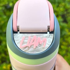 Peut inclure: Gros plan sur une gourde multicolore avec le nom "LIAM" en lettres roses. La gourde présente un couvercle rose, une bande bleu-vert, une bande vert clair et une base rose et bleu clair. Le fond est vert flou.