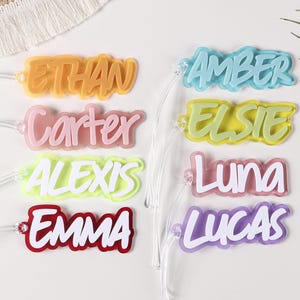 May include: A collection of colorful acrylic name tags with white lettering. Names include Ethan, Amber, Carter, Elsie, Alexis, Luna, Emma, and Lucas. Each tag has a clear string attached for easy use. The tags are in various colors, including orange, blue, pink, yellow, red, and purple.