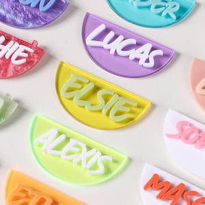 May include: A selection of colourful, semi-circular name tags with white, cursive lettering. The tags are in various colours, including red, purple, yellow, and green. Names like "Sophie", "Lucas", and "Elsie" are visible. The tags are arranged on a white surface.