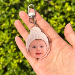 Op de afbeelding: Transparante acryl sleutelhanger met een lachende baby met een witte gebreide muts. De sleutelhanger heeft een zilverkleurige metalen sluiting en ring. De achtergrond is een vage groene blad.