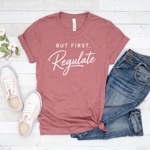 May include: A dusty rose t-shirt with the text "BUT FIRST, Regulate" in white script. The shirt is paired with blue jeans and white trainers. The t-shirt is folded at the sleeves.