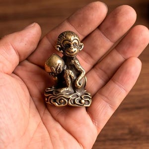 Könnte beinhalten: Eine kleine, metallische, messingfarbene Affenfigur, die auf einem stilisierten Wolkensockel sitzt. Der Affe hält einen runden, pfirsichförmigen Gegenstand in seiner rechten Hand. Die Oberfläche der Figur weist detaillierte, texturierte Gravuren auf.