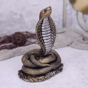 Puede incluir: Una figura de cobra enroscada con un cuerpo texturizado de color bronce y una capucha prominente y elevada. La parte inferior de la capucha muestra una serie de escamas horizontales lisas de tono plateado, con la cabeza inclinada hacia arriba.