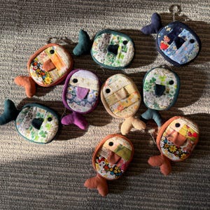 May include: A collection of handmade fish-shaped keychains. Each keychain features a patchwork design with various fabric patterns and colors, including floral, geometric, and solid hues. The fish have dark eyes, fins, and tail accents.