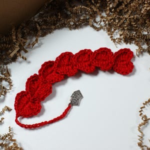 May include: A handmade red crochet bookmark with a series of heart-shaped designs. A small silver charm with playing card symbols hangs from the end. The bookmark is displayed on a white surface with brown paper filler.