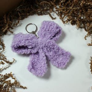 Può includere: Un portachiavi a forma di fiocco, soffice, color lavanda. Il portachiavi ha un anello color argento. Il fiocco è realizzato in un materiale morbido e strutturato. Lo sfondo è bianco con trucioli di carta marrone.