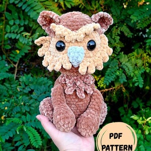 May include: A brown crocheted plush owl toy with a textured, fuzzy yarn finish. The owl features large black plastic eyes, a light blue beak, and light tan tufted eyebrows and facial feathers. A circular label with the text PDF PATTERN is visible.