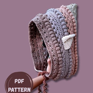 May include: A crocheted bonnet featuring a textured bobble stitch pattern in horizontal stripes of brown, mauve, and light gray. The bonnet includes a small, triangular cream-colored ear detail on the side and thin, curly crocheted ties at the base.