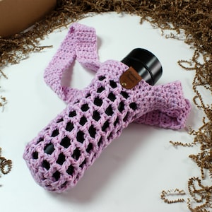May include: A light purple, crocheted water bottle holder with a strap. The holder has an open, net-like design and a small, brown leather tag. The holder is designed to fit a black water bottle. The strap is approximately 30 inches long.