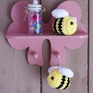 Puede incluir: Estante rosa en forma de lazo con un tarro de cristal lleno de flores de colores, una abeja de ganchillo amarilla y negra y un llavero de abeja. El estante tiene dos clavijas rosas y está montado en una pared de madera rosa claro.
