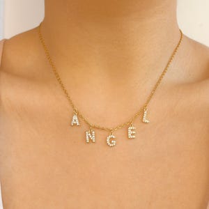 Peut inclure: Un collier doré avec le mot « ANGEL » épelé avec des pendentifs individuels en forme de lettres. Chaque lettre est incrustée de petits cristaux étincelants. Le collier est porté autour du cou.