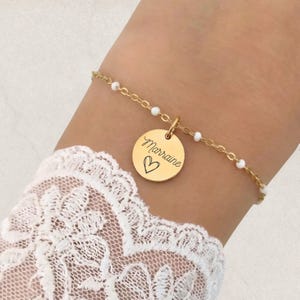 Peut inclure: Bracelet doré avec une chaîne délicate et de petites perles blanches. Un charm rond gravé du mot "Marrine" et d'un cœur est suspendu au bracelet. Le bracelet est porté au poignet, avec une manche en dentelle blanche visible.