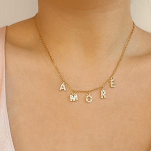 May include: A gold-colored necklace with the word "AMORE" spelled out in individual, sparkling letter pendants. The letters are encrusted with small, clear crystals, and the necklace is worn around the neck.