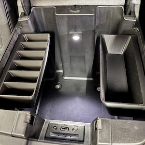 May include: Black car console with multiple storage compartments. Features a tray with dividers, a deep bin, and a central area with a light. Includes USB and AUX ports.