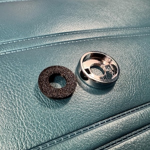 May include: A chrome-finished metal component and a black, circular rubber washer rest on a teal, textured surface. The metal piece has a hollow center and a shiny, reflective surface. The rubber washer is dark and porous.