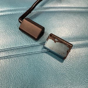 May include: Two rectangular, black and teal-colored car seat belt buckles. One buckle is black and attached to a black wire. The other buckle is teal with chipped paint. The background is a teal-colored, textured surface.