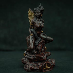 Puede incluir: Una figura de color marrón oscuro con cuernos y una estructura en forma de abanico detrás de la cabeza, sentada sobre una base rocosa. La figura presenta detalles dorados en los cuernos, joyas y un pequeño símbolo en la base. Una serpiente oscura se enrosca.