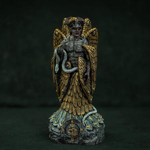 Puede incluir: Una figura texturizada de tonos oscuros que representa a un ser alado con cuernos que sostiene una serpiente enroscada. La figura presenta alas superpuestas de color oro y bronce, un torso musculoso y una base con un emblema circular.