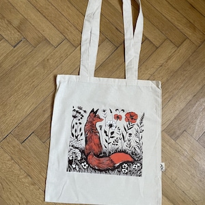 Pode incluir: Uma sacola de lona na cor creme com uma estampa gráfica central de uma raposa vermelha sentada entre flores silvestres e papoulas estilizadas em preto e branco. A bolsa possui duas alças longas e uma pequena etiqueta de tecido branca.
