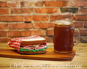 May include: A sandwich wrapped in a red and white checkered cloth, with layers of ham, cheese, and lettuce, sits on a wooden cutting board. A mason jar filled with a dark beverage is next to the sandwich. The background is a brick wall.