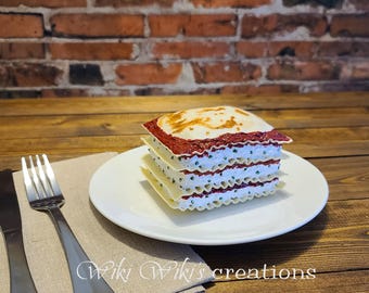 May include: A square serving of lasagna on a white plate, featuring alternating layers of wavy pasta, red tomato sauce, and a white cheese filling with green herbs. A silver fork and knife rest on a beige napkin beside the plate.