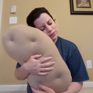May include: A person is hugging a large, beige, plush potato-shaped pillow. The pillow has a textured surface and is shaped like a potato.
