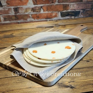 May include: A stack of four white tortillas with orange spots, wrapped in a light gray linen cloth, on a wooden cutting board.