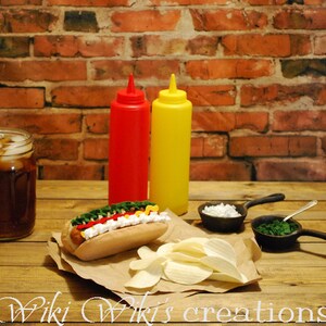 May include: A hot dog with ketchup, mustard, and relish on a white bun, next to a pile of potato chips, a glass of iced tea, two condiment bottles, and two small bowls with white and green toppings. The scene is set on a wooden table with a brick wall background.