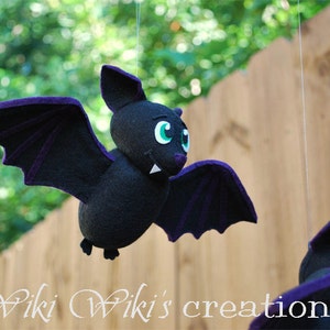 K&ouml;nnte beinhalten: Eine schwarze Filzfledermaus mit lila Fl&uuml;geln und gr&uuml;nen Augen h&auml;ngt an einer Schnur. Die Fledermaus hat ein zahniges Grinsen und ist eine niedliche Halloween-Dekoration.