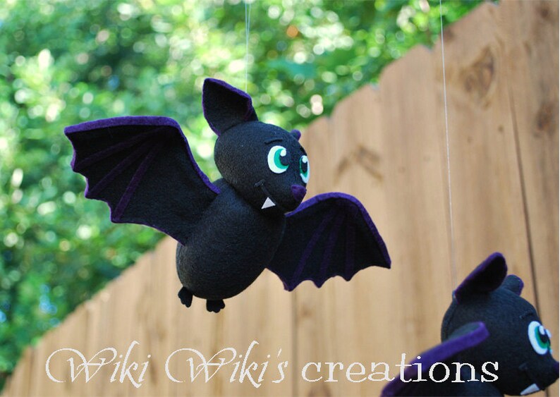 K&ouml;nnte beinhalten: Eine schwarze Filzfledermaus mit lila Fl&uuml;geln und gr&uuml;nen Augen h&auml;ngt an einer Schnur. Die Fledermaus hat ein zahniges Grinsen und ist eine niedliche Halloween-Dekoration.