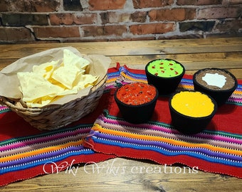 May include: A set of four felt food bowls filled with different dips, including guacamole, salsa, cheese, and sour cream. A basket of tortilla chips sits next to the bowls on a colorful striped placemat.