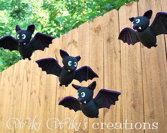 May include: Four black felt bats with purple accents hang from a wooden fence. Each bat has a cute, cartoon-like face with green eyes.