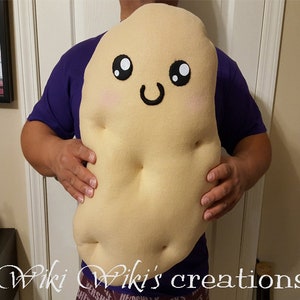 May include: A large, beige plush potato with a smiling face. The potato has black eyes, a black mouth, and pink cheeks. The potato is being held by a person.